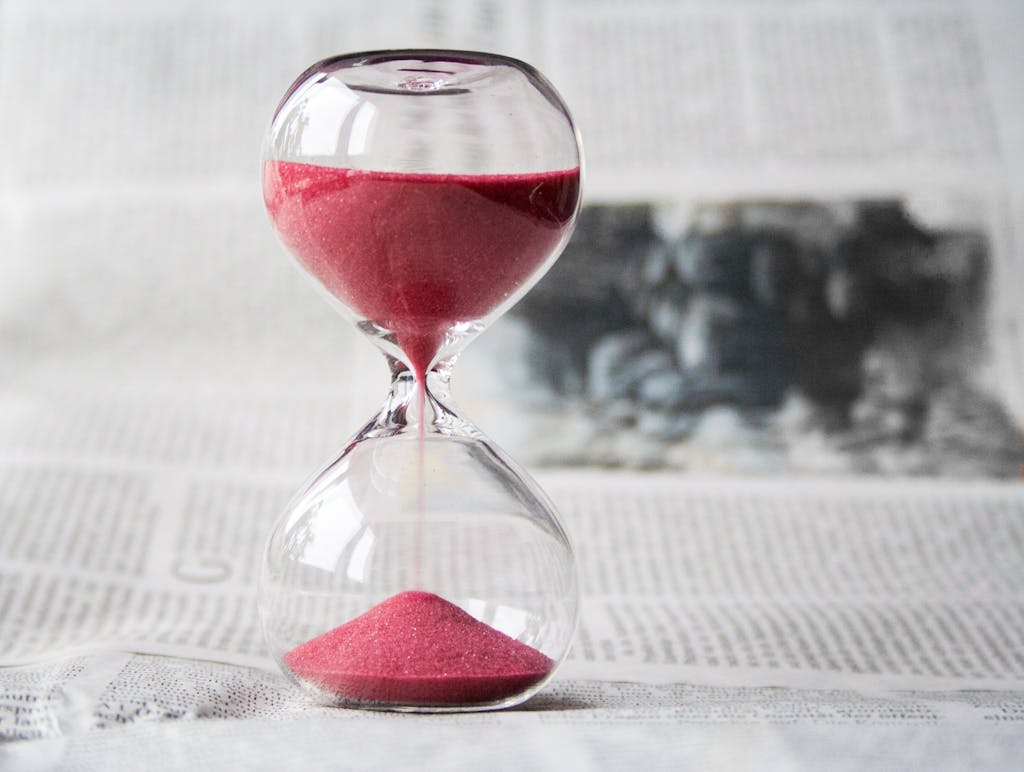 BLOG VIEW Naijastudyhub Close-up of a transparent hourglass with pink sand flowing, placed on a newspaper background.