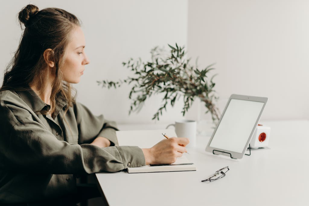 BLOG VIEW Naijastudyhub Freelancer writing notes in a minimalist home office with a white tablet, plant, and reading glasses.